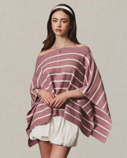 A woman modeling an off-the-shoulder knit poncho sweater with horizontal dusty rose and white stripes, paired with white shorts, against a plain light beige background.