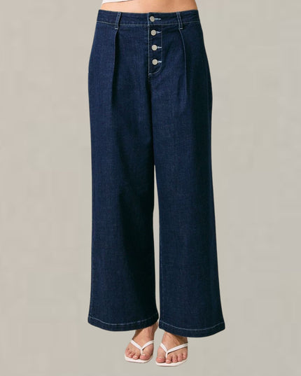 A person wearing dark blue wide-leg denim pants with a high waist, front pleats, and a button fly, shown from the waist down against a plain light gray studio background.