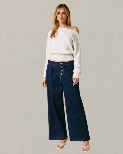 A woman modeling a white off-the-shoulder cropped knit top and dark blue wide-leg denim jeans with front buttons, paired with white heeled sandals, against a plain light gray studio background.