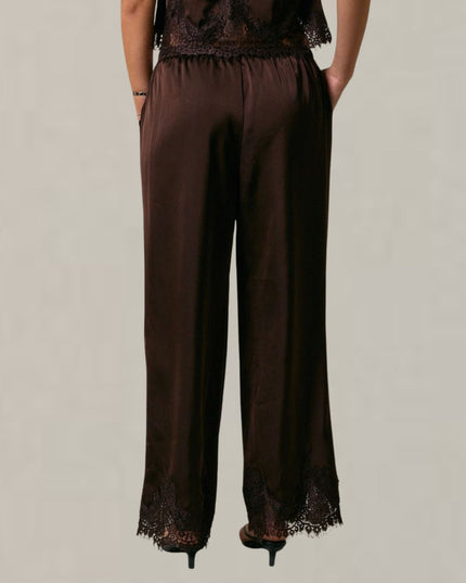 A person shown from the back wearing dark brown wide-leg pants with black lace trim at the waistband and hem, paired with black heels against a plain light background.