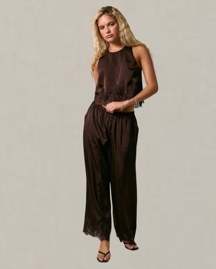 A woman modeling a dark brown sleeveless jumpsuit with lace trim details, standing against a neutral light gray studio background.