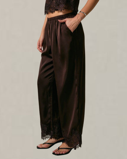A model wearing dark brown wide-leg satin pants with lace trim at the hem, paired with black sandals and a matching dark lace top, shown against a plain light gray background.