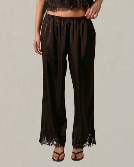 A model is wearing dark brown wide-leg pants with black lace trim at the hem, paired with black sandals, against a plain light gray background.