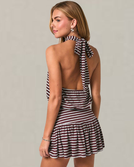 A woman modeling a sleeveless halter mini dress from the back, featuring a black and white horizontal stripe pattern and a ruffled tie detail at the back of the neck, set against a plain light gray studio background.