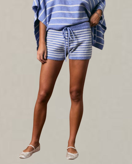 A model wearing a matching knit lounge set consisting of a blue and white striped poncho and shorts with a drawstring waist, paired with white ballet flats, against a solid light gray background.