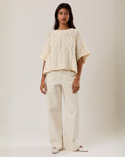 Woman wearing a cream-colored cable knit sweater and wide-leg pants on a plain background