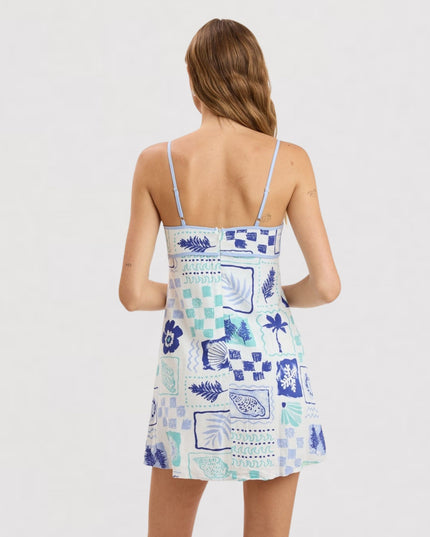 A woman shown from the back wearing a sleeveless spaghetti-strap dress with a blue and white tropical patchwork print, set against a plain light gray studio background.