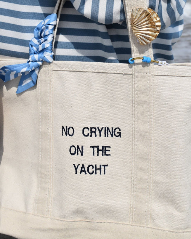 A large off-white canvas tote bag with black embroidered text reading 'NO CRYING ON THE YACHT'. The bag has a blue-and-white striped fabric handle wrap tied in a bow, a gold seashell-shaped charm, and a blue carabiner.