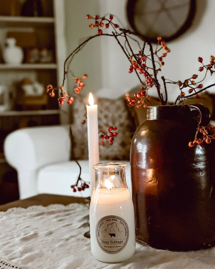Candle in a bottle with a label on a table with a vase of red berries in a cozy living room.