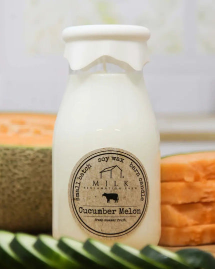 Candle in a milk bottle with a label, surrounded by cucumbers and melons on a wooden surface.
