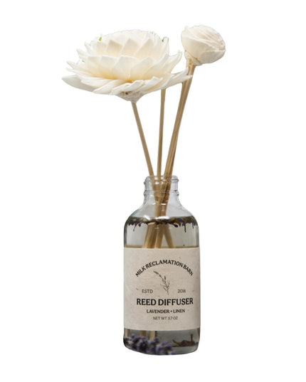 A clear glass reed diffuser bottle with a cream label reading 'Her Reclamation Wash' and 'Reed Diffuser Lavender + Linen,' holding several natural reed sticks with off-white flower heads, shown on a plain white background.