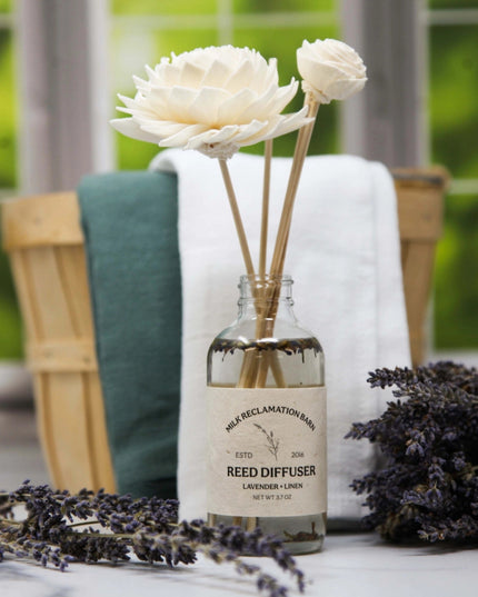 A clear glass reed diffuser bottle with a cream label reading 'Highland Declamation Born' and 'Reed Diffuser Lavender + Lin,' filled with water and holding two dried flower stems. The bottle is styled on a tabletop with lavender sprigs, a white towel, a teal towel, and a wooden basket in front of a softly blurred window.