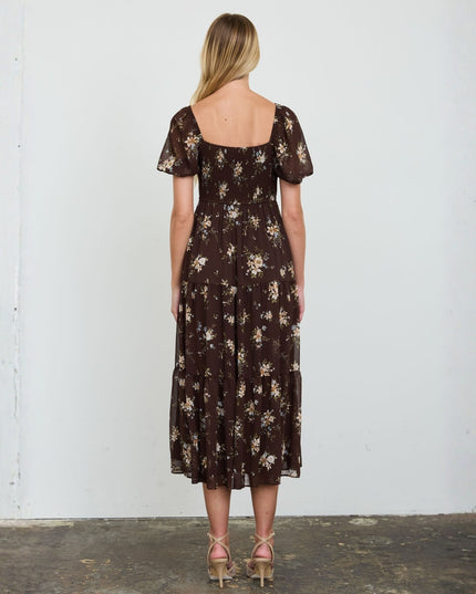 A woman stands with her back to the camera wearing a dark brown midi dress with a beige and white floral pattern, short puff sleeves, and a square neckline. She is standing in front of a plain white wall.