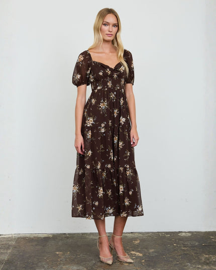 A woman stands against a plain white wall wearing a dark brown midi dress with a beige and white floral print, short puff sleeves, and a square neckline. She is wearing beige high-heeled sandals.