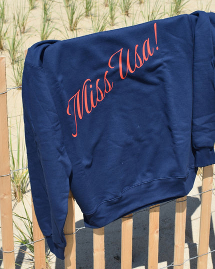 Blue sweatshirt with 'Miss USA!' text on a wooden fence by a sandy beach.
