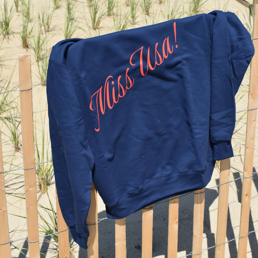 Blue sweatshirt with 'Miss USA!' text on a wooden fence by a sandy beach.