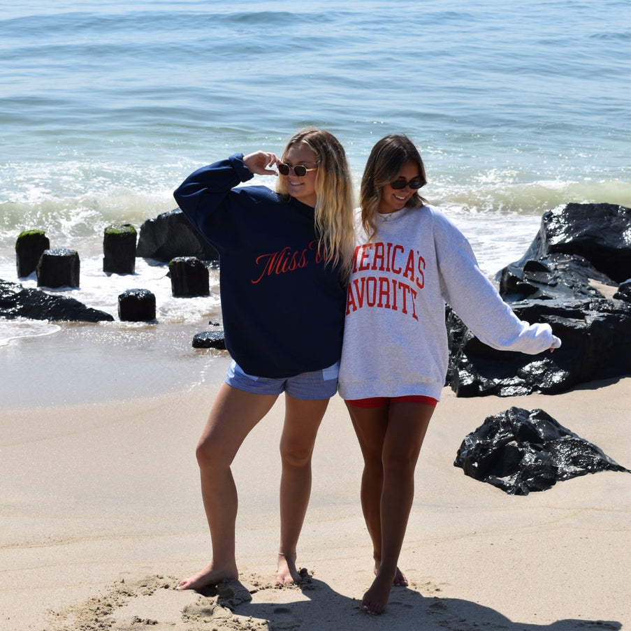 Two women standing on a beach wearing oversized sweatshirts with visible text.