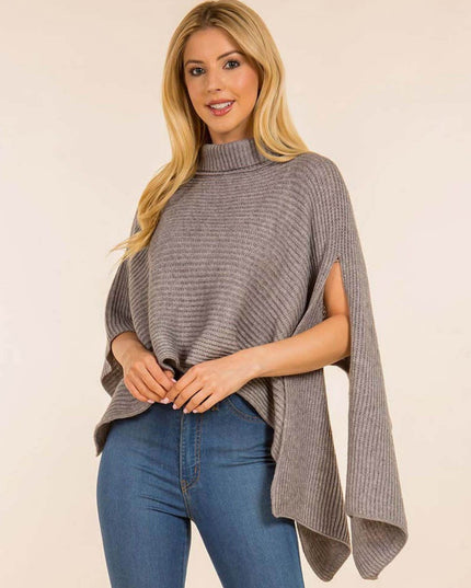 A blonde woman wearing a gray ribbed knit poncho sweater with side cutouts and blue jeans, standing against a plain light beige studio background.