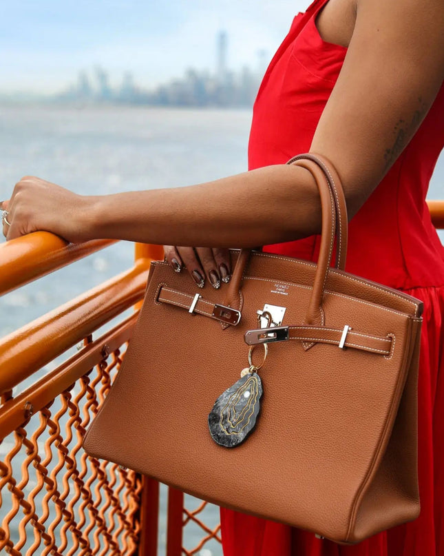 A woman in a red dress holds a tan leather handbag with silver hardware and a hanging stone charm, standing by an orange railing with water and a blurred city skyline in the background.