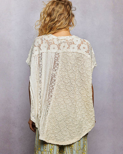 A woman viewed from behind wearing a cream-colored, textured lace and crochet patchwork top with short sleeves, paired with patterned green and yellow pants, set against a plain gray studio background.