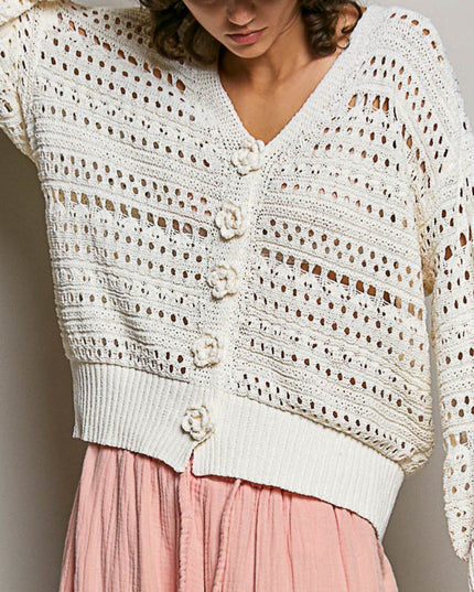 A close-up of a person wearing a cream open-knit cardigan with large decorative buttons and a pink skirt against a plain light gray background.