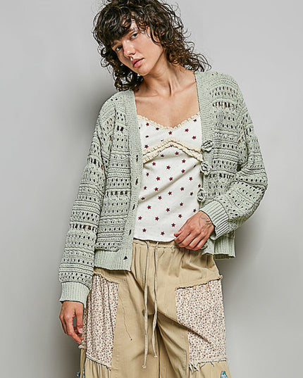 A model wearing a sage green open-knit cardigan over a white lace-trim camisole with red polka dots, paired with beige patchwork cargo pants, against a solid gray studio background.