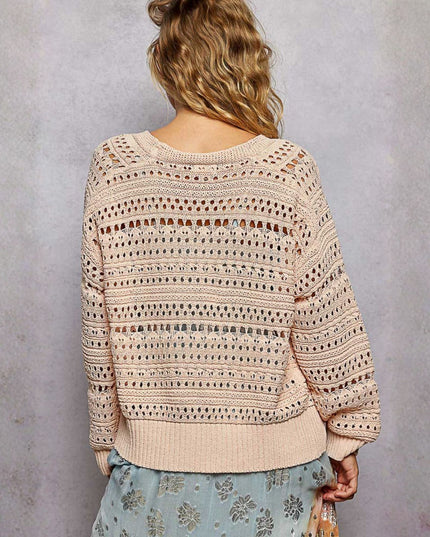 A woman viewed from behind wearing a cream-colored open-knit sweater with horizontal striped patterns and a ribbed hem, paired with a light blue floral skirt, set against a textured gray wall.