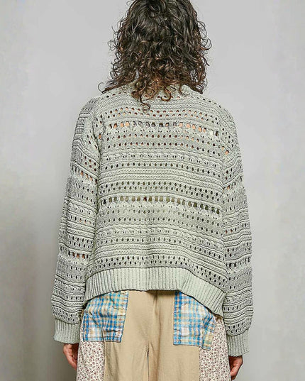 A person shown from the back wearing a light sage green open-knit sweater with geometric patterns, paired with beige pants featuring blue patch pockets, against a neutral gray background.