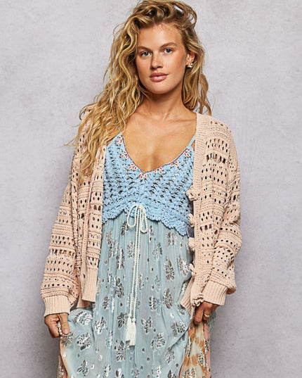 A woman modeling a light blue crochet crop top and a beige open-knit cardigan over a light blue floral maxi dress, set against a textured gray studio background.