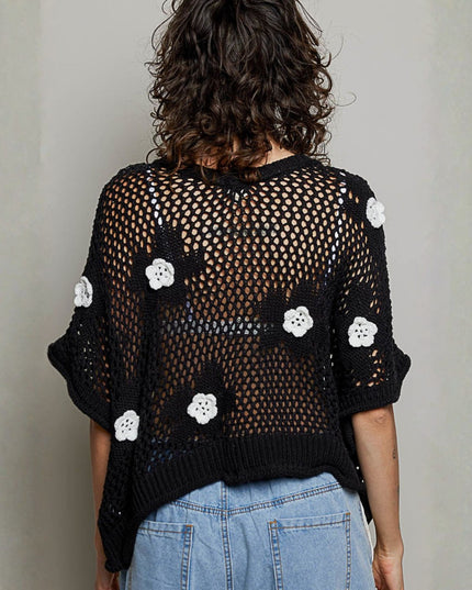 A woman viewed from behind wearing a black open-knit cropped sweater with white crocheted flower appliqués, paired with light blue jeans, against a plain light gray wall.