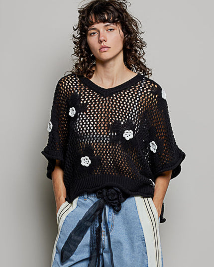 A model wearing a black open-knit V-neck sweater with white flower appliqués, paired with light blue jeans featuring white side stripes, against a solid gray studio background.