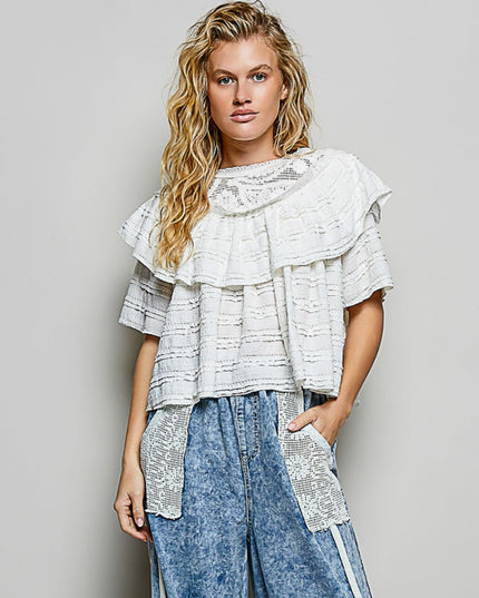 A woman modeling a white ruffled short-sleeve top with textured horizontal stripes and blue denim wide-leg pants with lace side panels, against a solid gray studio background.