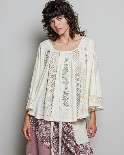 A woman modeling a cream-colored bohemian-style top with floral embroidery and lace details, paired with patterned pants, against a plain gray studio background.