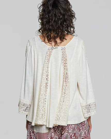 A woman shown from the back wearing a cream-colored, long-sleeved bohemian-style blouse with intricate lace trim down the center and on the cuffs, paired with patterned pink pants, against a solid gray background.