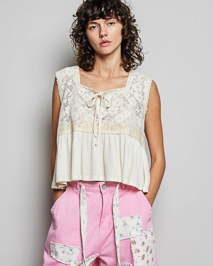 A model wearing a sleeveless cream lace and textured top with a tie neckline, paired with pink patchwork pants, against a plain light gray background.