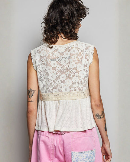 A woman viewed from behind wearing a sleeveless cream lace top with a peplum hem and pink pants, standing against a plain light gray wall.