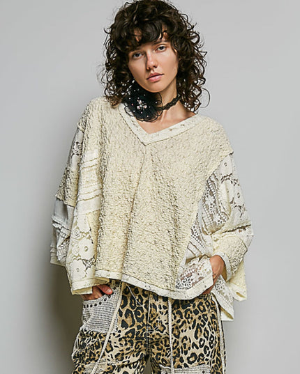 A model with curly dark hair wearing a cream-colored, textured patchwork knit top with lace sleeves and leopard-print pants, standing against a plain light gray background.