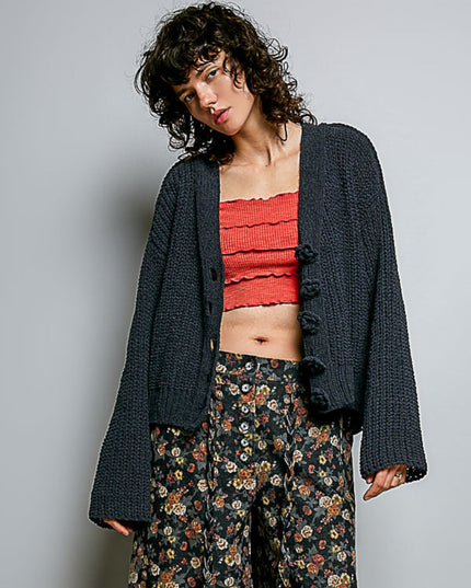 A model wearing a dark charcoal gray chunky knit cardigan with pom-pom buttons over a red ruffled tube top and dark floral wide-leg pants, set against a plain light gray studio background.