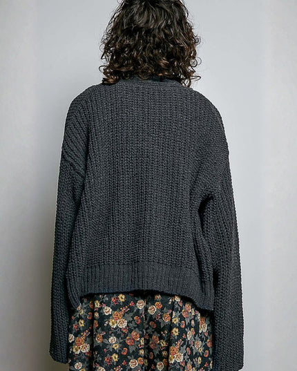 A person shown from the back wearing a dark charcoal gray chunky knit cardigan and a dark floral skirt, standing against a plain light gray wall.