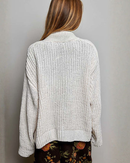 A woman viewed from behind wearing a cream-colored, textured open-knit sweater and dark floral pants against a plain gray wall.