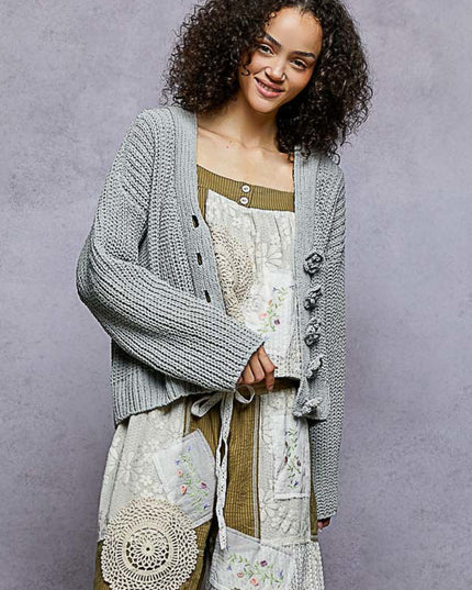 A woman with curly dark hair models a light gray chunky knit cardigan over a cream patchwork dress with olive green trim, set against a textured gray studio background.