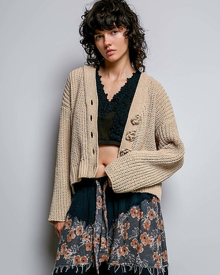 A model wearing a beige chunky knit cardigan with decorative buttons over a black lace top and a dark floral skirt, photographed against a neutral gray studio background.