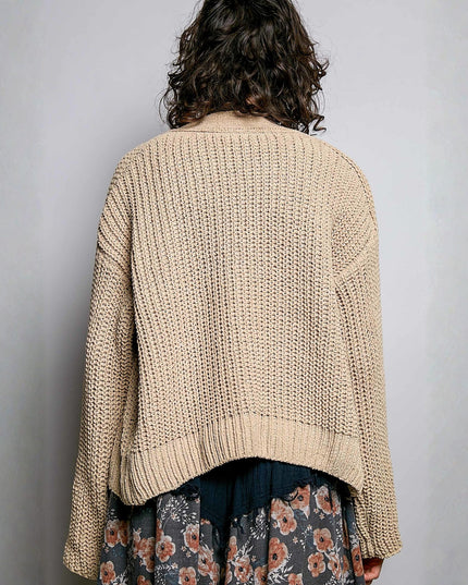 A person shown from the back wearing a chunky knit cropped cardigan in a light beige color, paired with a dark skirt featuring a floral pattern. The background is a plain light gray wall.