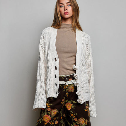 A woman models a cream-colored, chunky open-front cardigan with decorative knotted buttons, worn over a beige mock-neck top and dark brown floral wide-leg pants, set against a plain gray studio background.