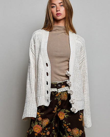 A woman models a cream-colored, chunky open-front cardigan with decorative knotted buttons, worn over a beige mock-neck top and dark brown floral wide-leg pants, set against a plain gray studio background.