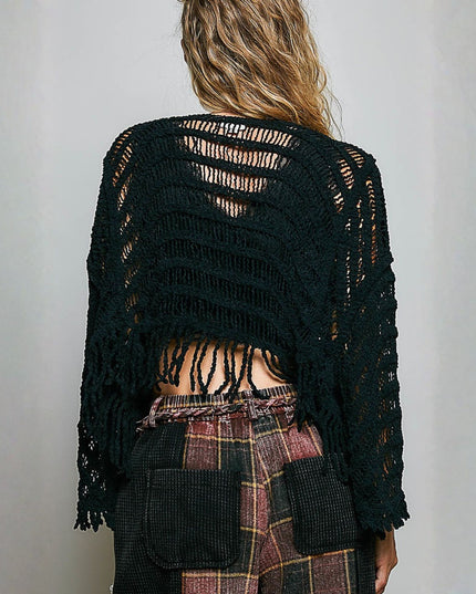 A woman viewed from behind wearing a black open-knit cropped cardigan with fringe, paired with plaid pants, against a plain light gray wall.
