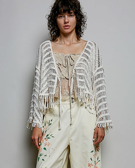 A model wearing a cream-colored crochet cardigan with fringe over a beige lace top and cream floral pants, standing against a solid gray studio background.