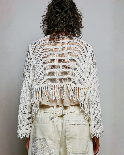 A person shown from the back wearing a cream-colored, open-knit cropped cardigan with fringe trim over a light beige top and cream pants, set against a plain light gray wall.