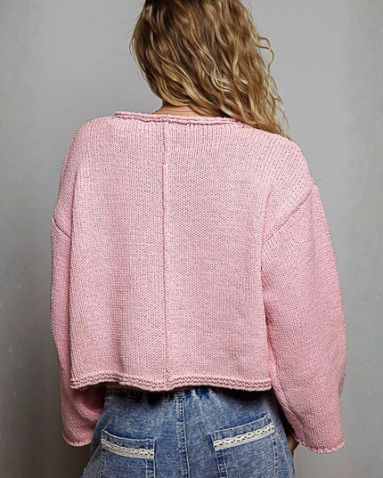 A woman viewed from behind wearing a light pink cropped knit sweater and blue jeans with white embroidery on the back pockets, set against a plain gray wall.