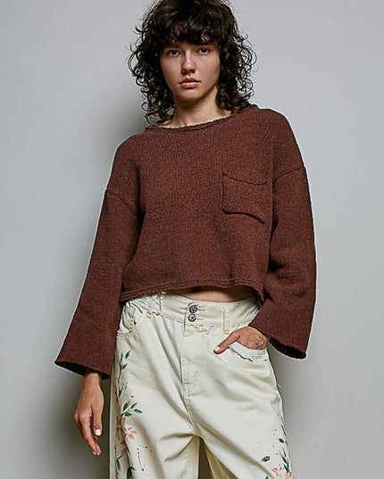 A model wearing a cropped, long-sleeve knit sweater in a heathered brownish-red color, paired with cream-colored floral pants, against a plain gray studio background.